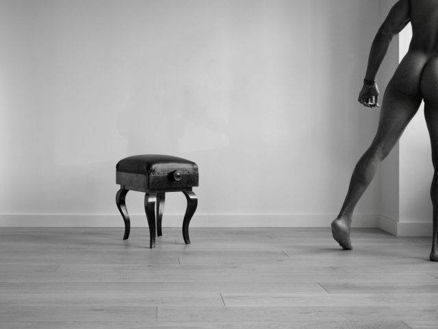 A black and white photograph of a nude man walking away from a piano bench.