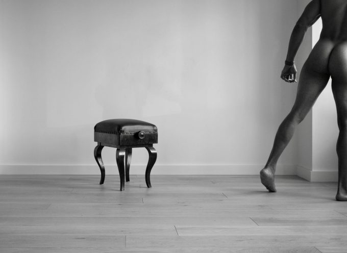 A black and white photograph of a nude man walking away from a piano bench.