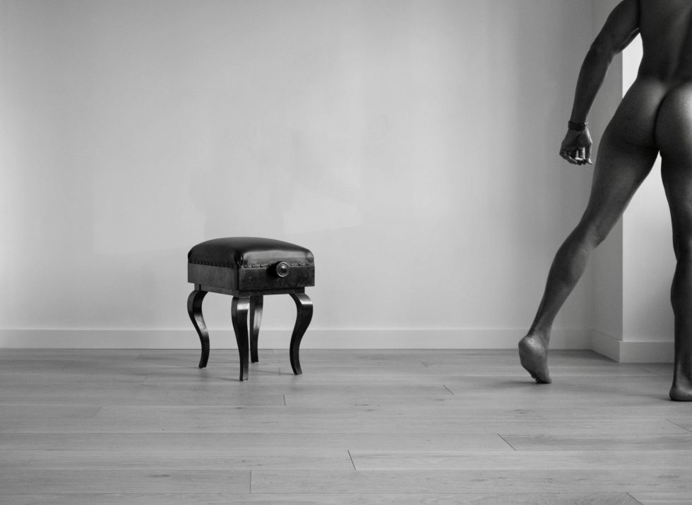 A black and white photograph of a nude man walking away from a piano bench.
