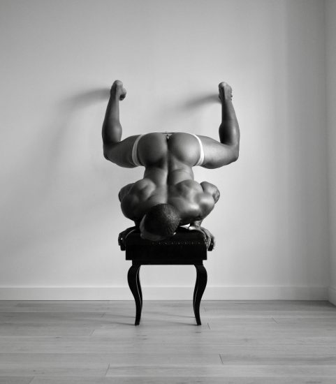 A black and white photograph of a nude man upside down on his head on a piano bench.