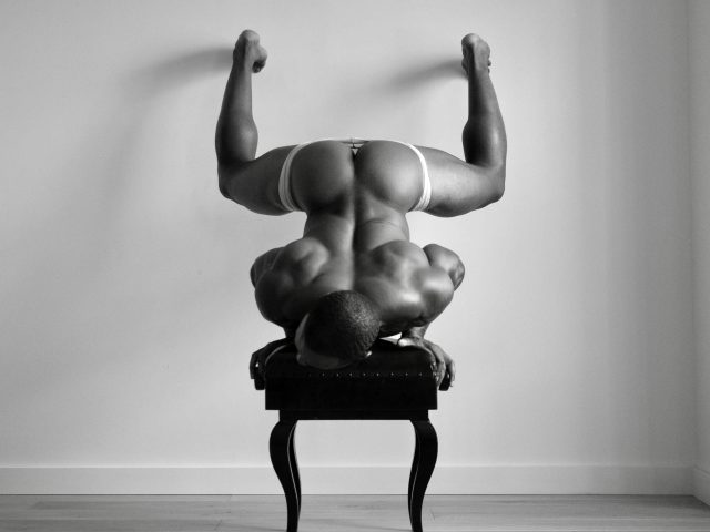 A black and white photograph of a nude man upside down on his head on a piano bench.