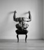 A black and white photograph of a nude man upside down on his head on a piano bench.