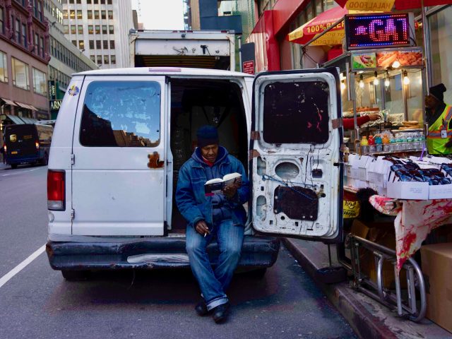 A color photograph of a man reading a book in the back of a white van.