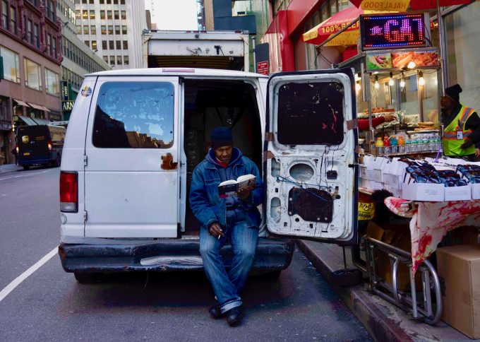 A color photograph of a man reading a book in the back of a white van.