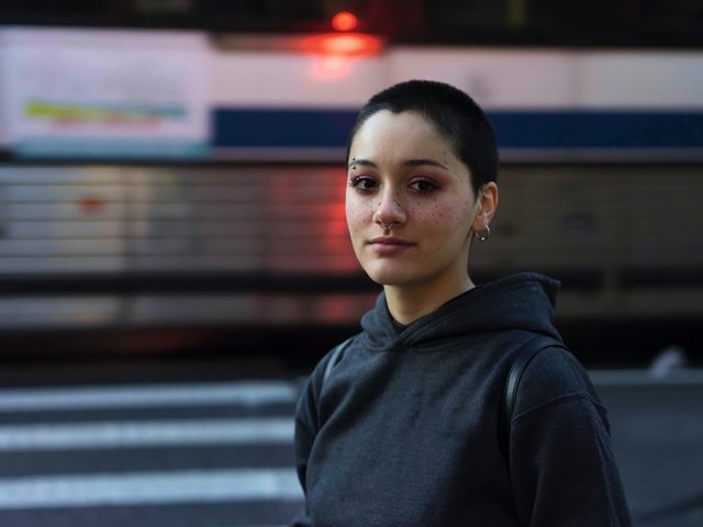 A color photograph of a striking young woman with freckles and short hair in front of a bus.