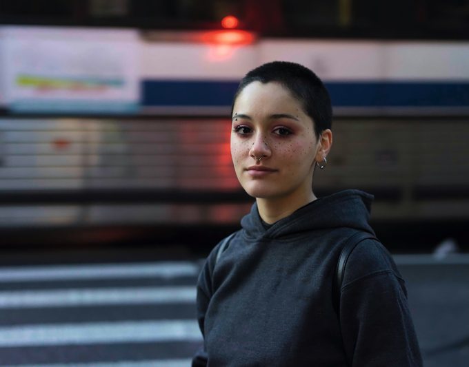 A color photograph of a striking young woman with freckles and short hair in front of a bus.