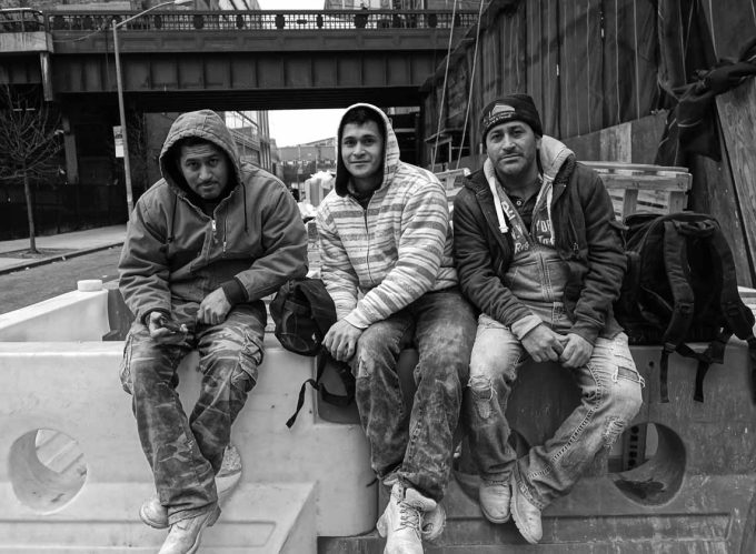 A black and white photograph of three men sitting on a barricade at the end of the work day.