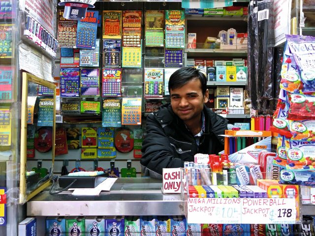 A photograph of a man in a newsstand smiling in front of lottery tickets.