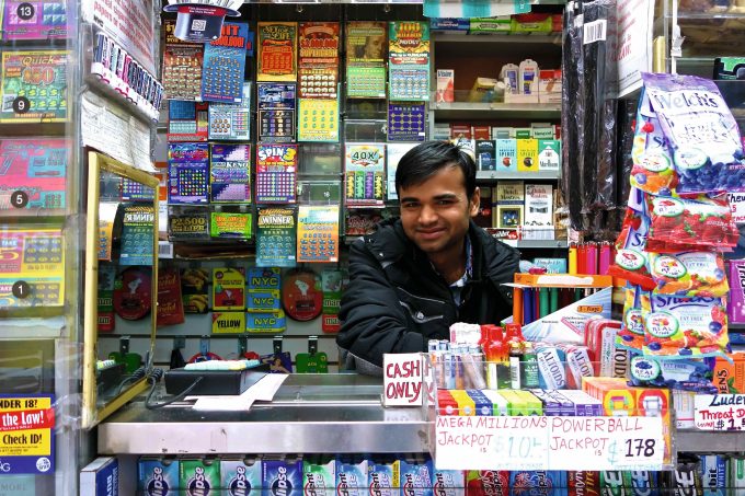 A photograph of a man in a newsstand smiling in front of lottery tickets.