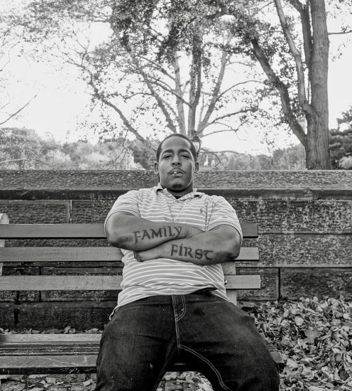 A black and white photograph of a man sitting on a park bench with his arms crossed.