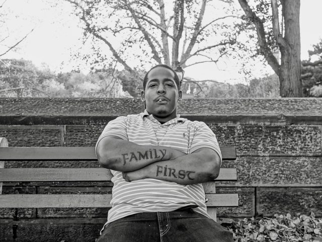 A black and white photograph of a man sitting on a park bench with his arms crossed.