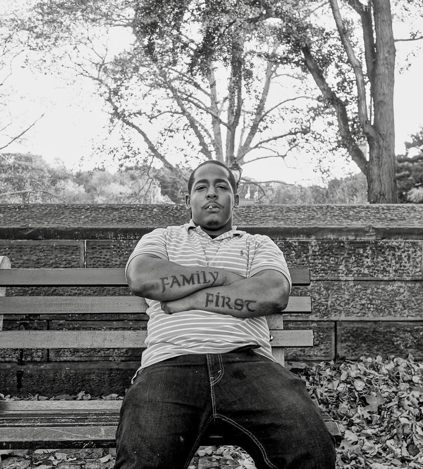 A black and white photograph of a man sitting on a park bench with his arms crossed.