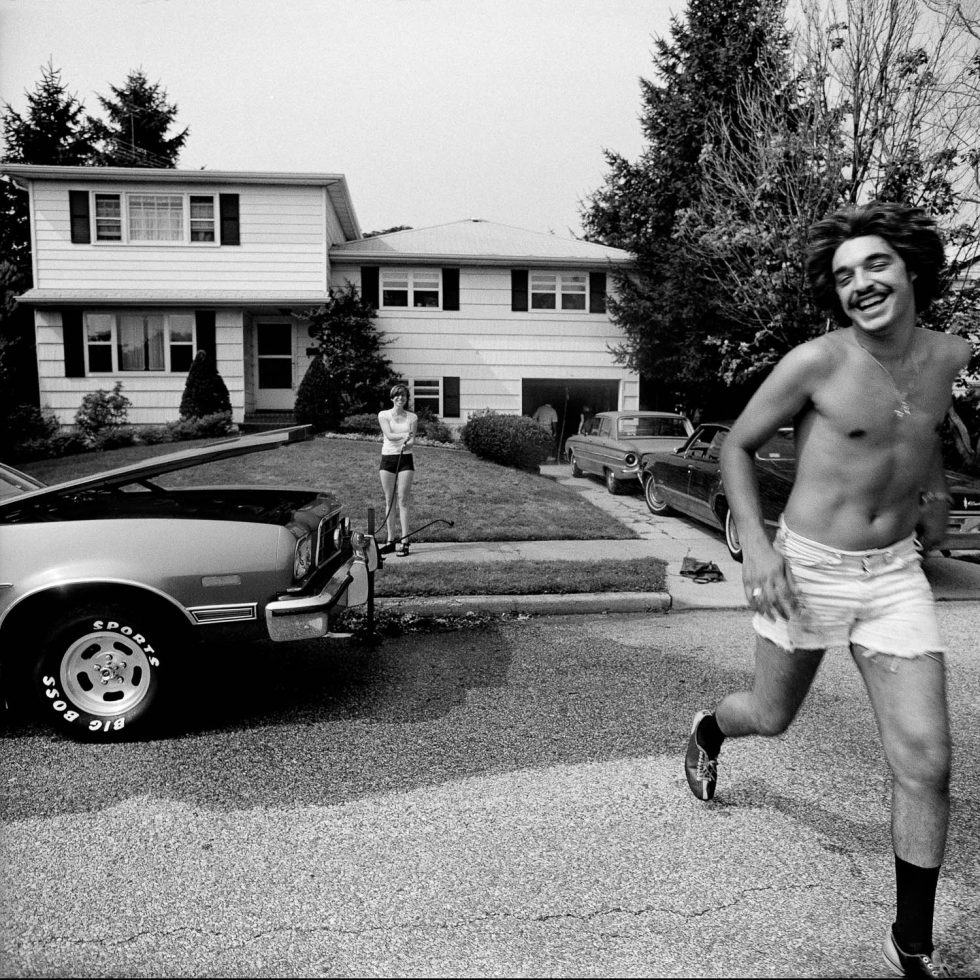 A black and white photograph of a shirtless young man running into a street in a suburban setting.