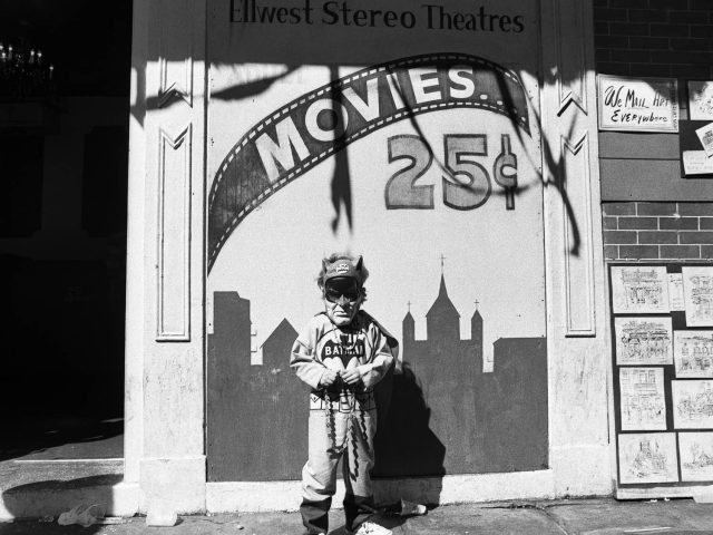 A black and white photograph of a young boy in costume standing in front of a movie theater sign.