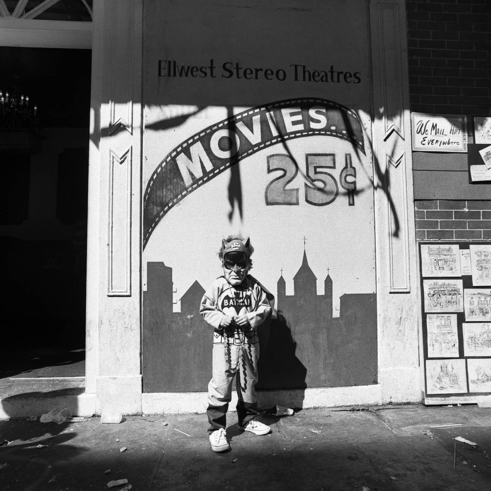 A black and white photograph of a young boy in costume standing in front of a movie theater sign.