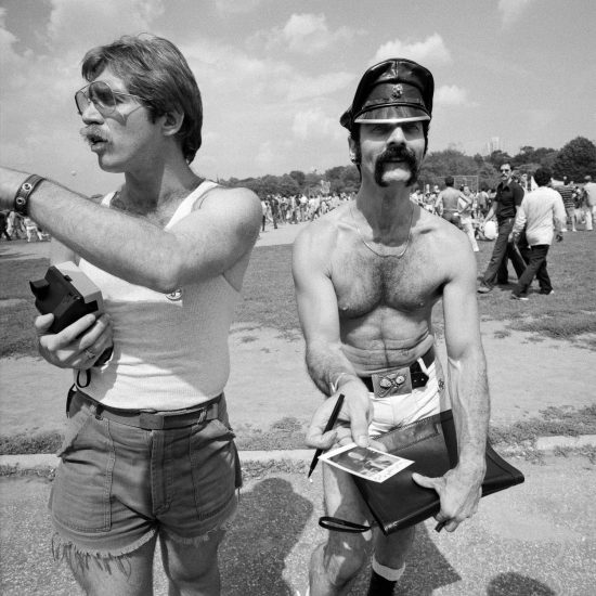 A black and white photograph of two gay men handing out polaroids.