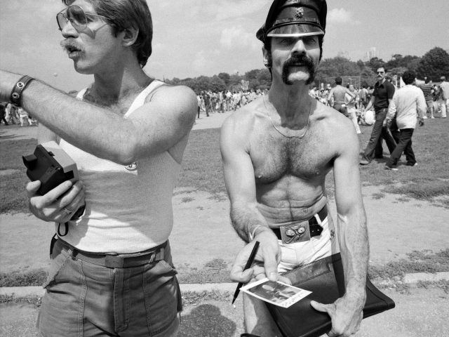 A black and white photograph of two gay men handing out polaroids.