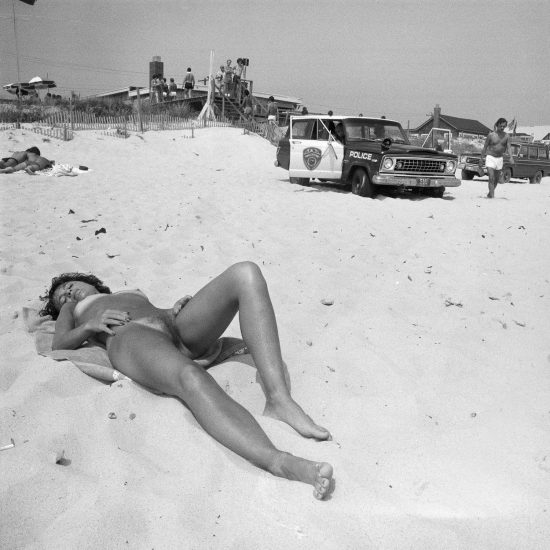 A black and white photograph of a woman suntanning on a beach next to a cop car.