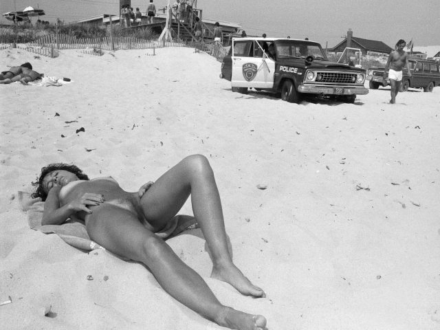 A black and white photograph of a woman suntanning on a beach next to a cop car.
