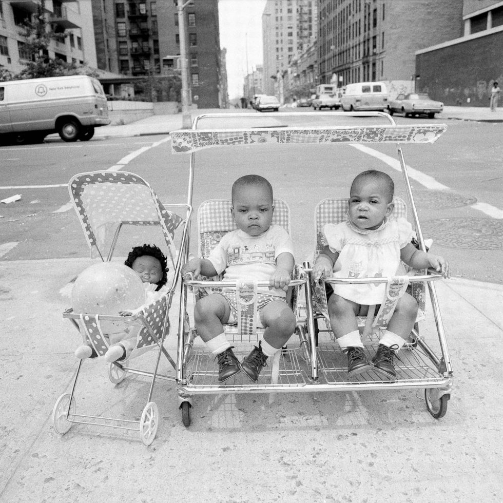 A black and white photograph of two babies in a double stroller with a doll in a stroller beside them.