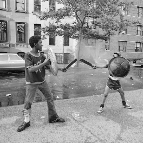 A black and white photograph of two boys playing with trash can lids and swords.