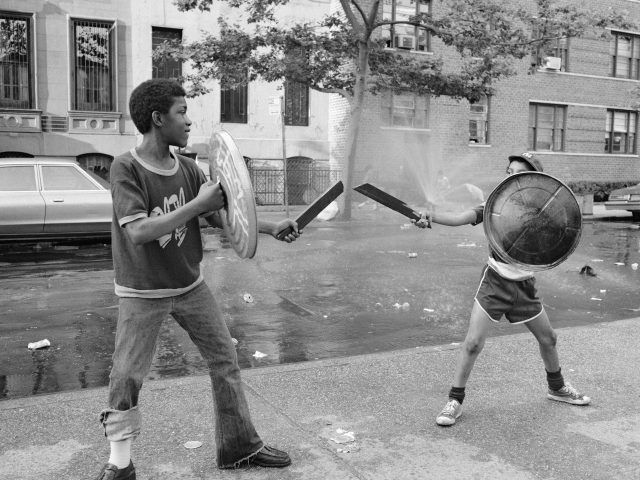A black and white photograph of two boys playing with trash can lids and swords.