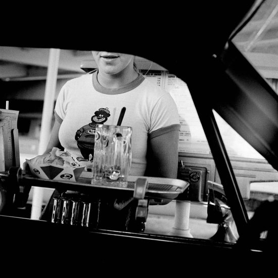 A black and white photograph of a young waitress bringing a tray of food to a passenger at a drive in window.