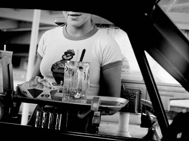 A black and white photograph of a young waitress bringing a tray of food to a passenger at a drive in window.