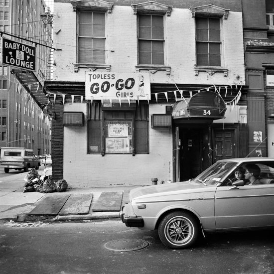 A black and white photograph of a car in front of a strip club.
