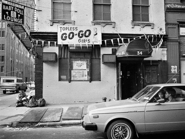 A black and white photograph of a car in front of a strip club.