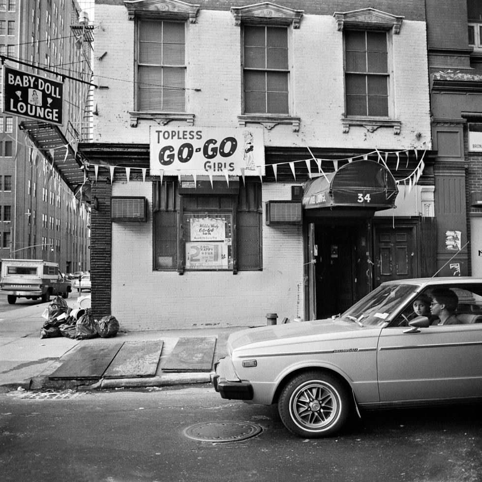 A black and white photograph of a car in front of a strip club.