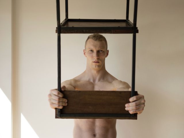 Shirtless person holding a wooden and metal chair frame in front of their chest against a neutral wall.
