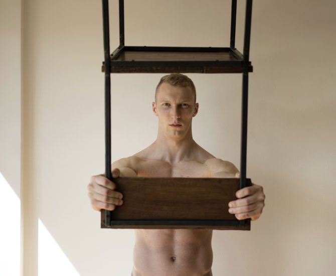 Shirtless person holding a wooden and metal chair frame in front of their chest against a neutral wall.