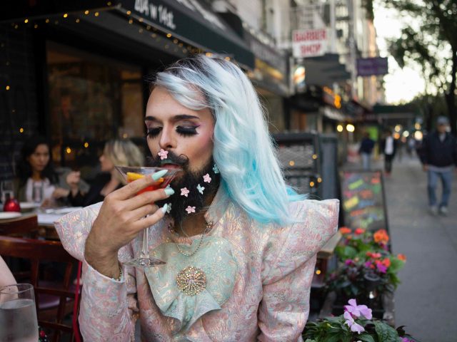 A color photograph of a drag queen drinking a cocktail al fresco on a city street.