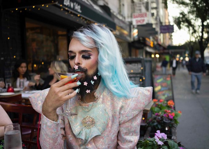 A color photograph of a drag queen drinking a cocktail al fresco on a city street.