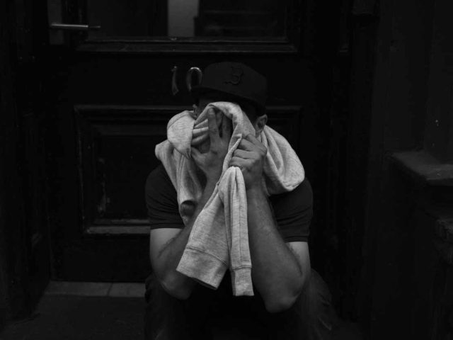 A black and white photograph of a man holding a sweater in front of his face in an alley.