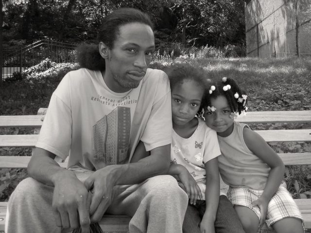 A black and white photograph of a young family sitting on a park bench.