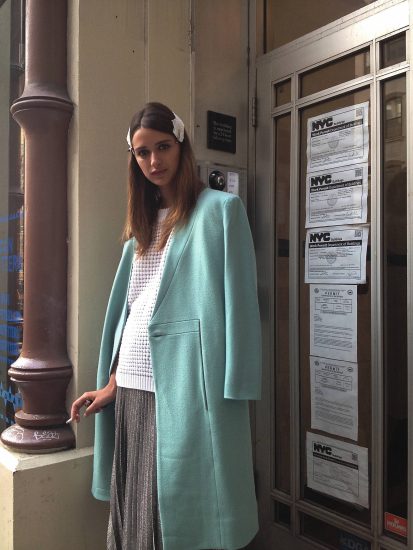 A color photograph of a young female model in a teal coat smoking a cigarette.