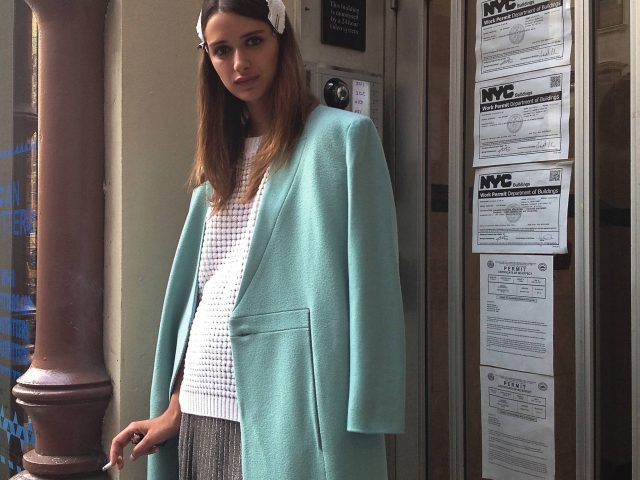 A color photograph of a young female model in a teal coat smoking a cigarette.