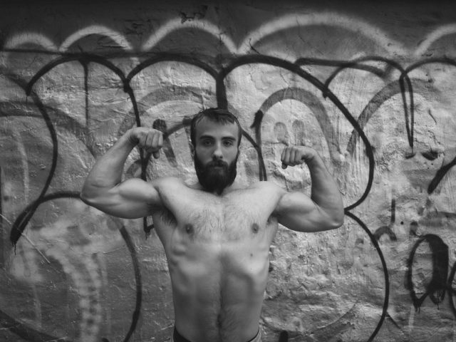 A black and white photograph of a strong young man flexing shirtless with a bushy beard.