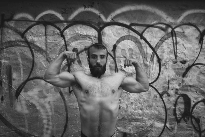 A black and white photograph of a strong young man flexing shirtless with a bushy beard.