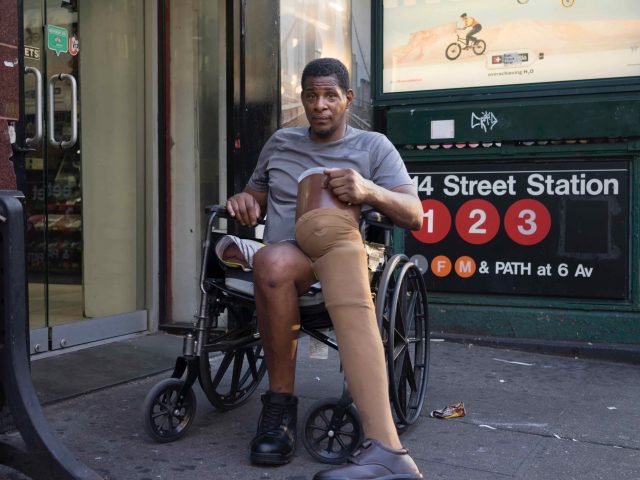 A color photograph of a man in a wheelchair with a prosthetic leg next to a subway entrance.