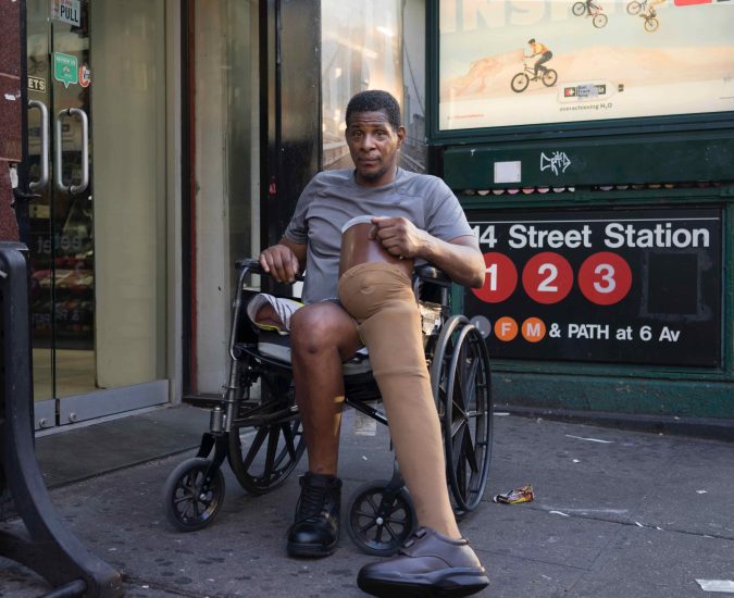 A color photograph of a man in a wheelchair with a prosthetic leg next to a subway entrance.