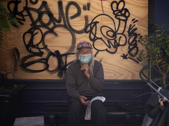 A color photograph of a man sitting in the shade in front of a graffiti covered piece of plywood.