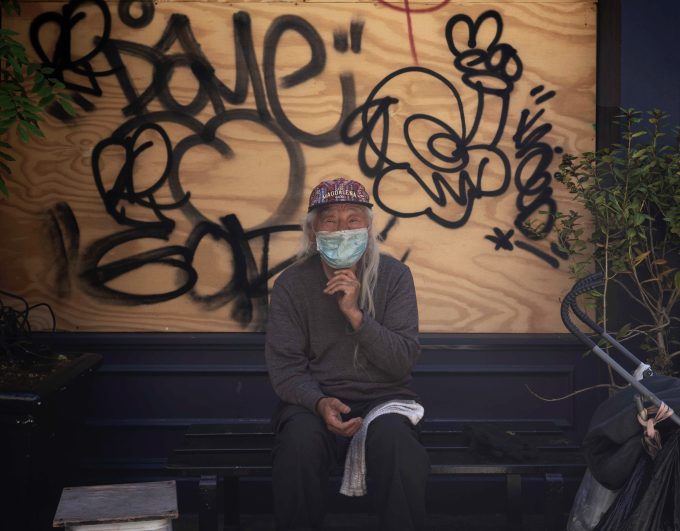 A color photograph of a man sitting in the shade in front of a graffiti covered piece of plywood.