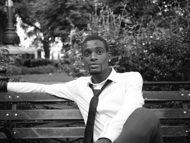 A black and white photograph of a young man on a park bench.