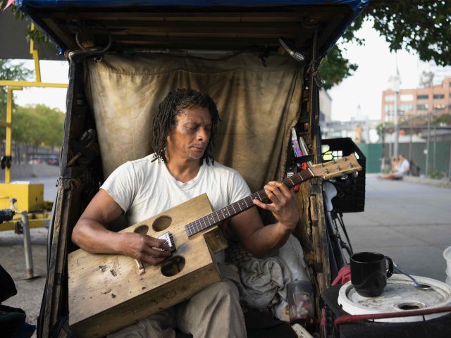 A color photograph of a man sitting under a makeshift shelter playing a handmade guitar.