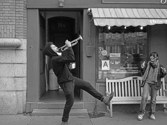 A black and white photograph of a man walking down the street blowing a trumpet.