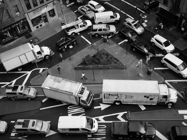 A black and white photograph of a variety of cars and trucks stuck in a traffic jam around a tiny triangular park photographed from above.