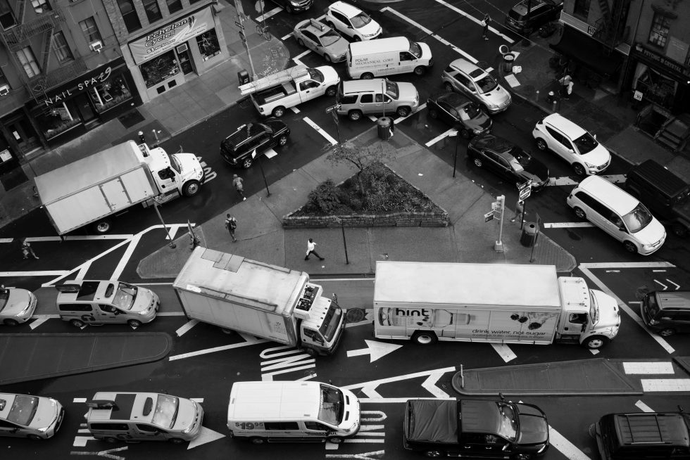 A black and white photograph of a variety of cars and trucks stuck in a traffic jam around a tiny triangular park photographed from above.
